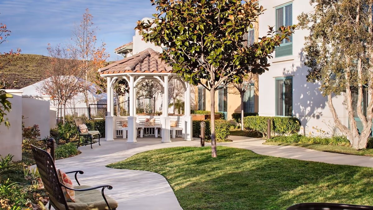 Outdoor garden area at Belmont Village Thousand Oaks featuring a white gazebo with a tiled roof, surrounded by benches and trees, with a paved walkway and green grass.
