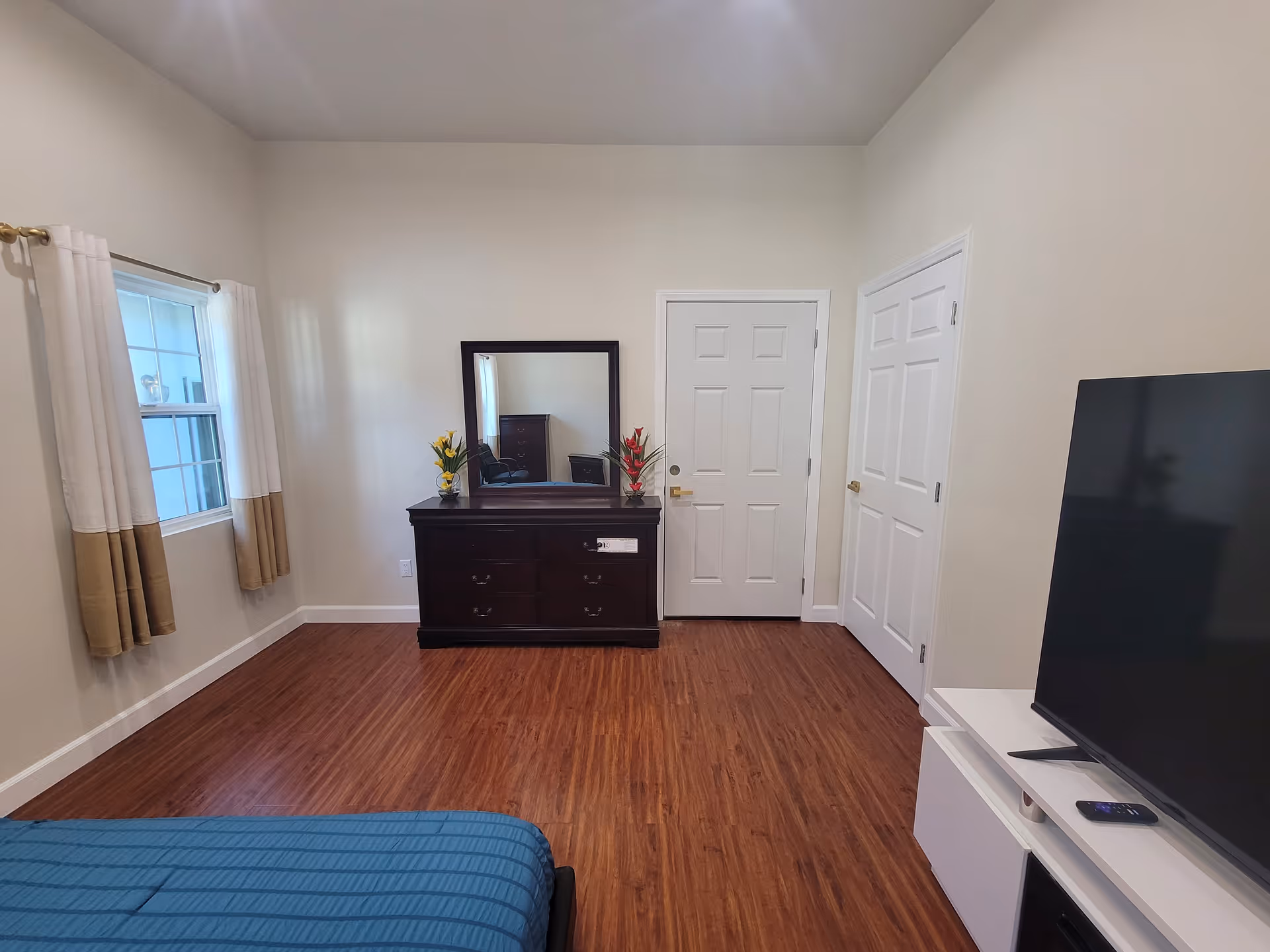 A bedroom with wooden flooring, a window with beige and white curtains on the left wall, a dark wooden dresser with a mirror and two flower vases, two white doors on the far wall, and a TV on a white stand on the right side. A blue bedspread is partially visible in the foreground.