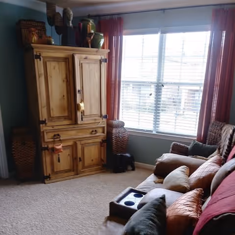 A cozy living room with a large wooden cabinet, a window with red curtains, and a comfortable sofa with multiple cushions. The room has carpeted flooring and decorative items on top of the cabinet.