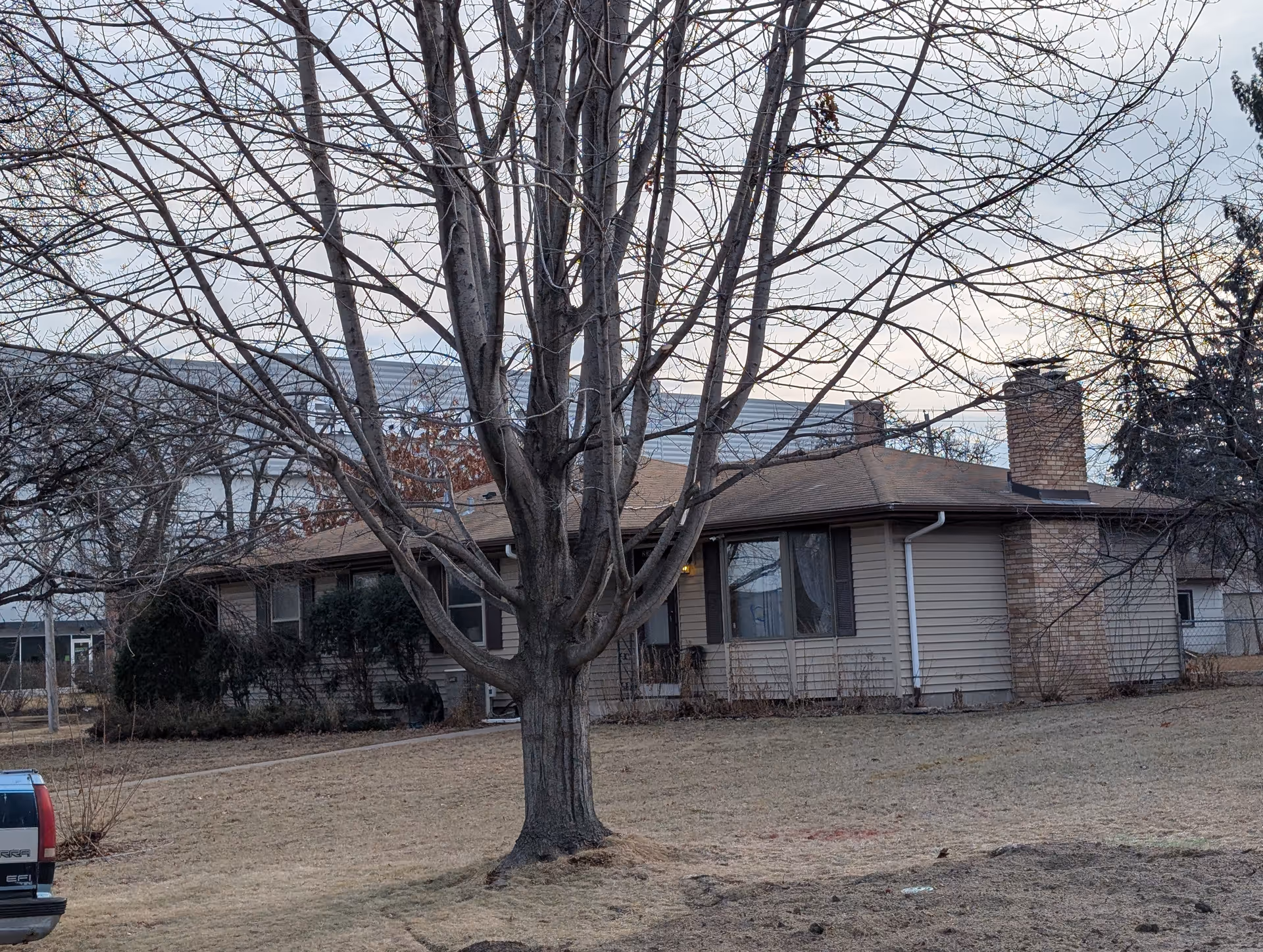 Single-story residential building with beige siding and a brown roof, featuring a brick chimney. In front of the building is a large leafless tree and a dry grassy yard. A red vehicle is partially visible on the left side of the image. The sky is cloudy and overcast.