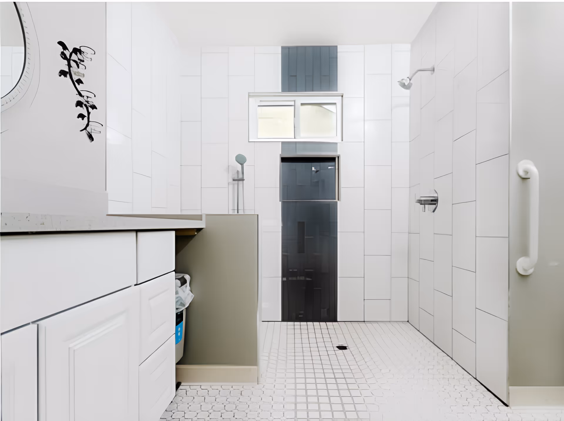A modern bathroom with white tiled walls and floor. There is a walk-in shower area with a showerhead on the right wall and a grab bar next to it. A small window is above the shower area, and there is a cabinet with white drawers and a countertop on the left side. The overall design is clean and minimalistic.