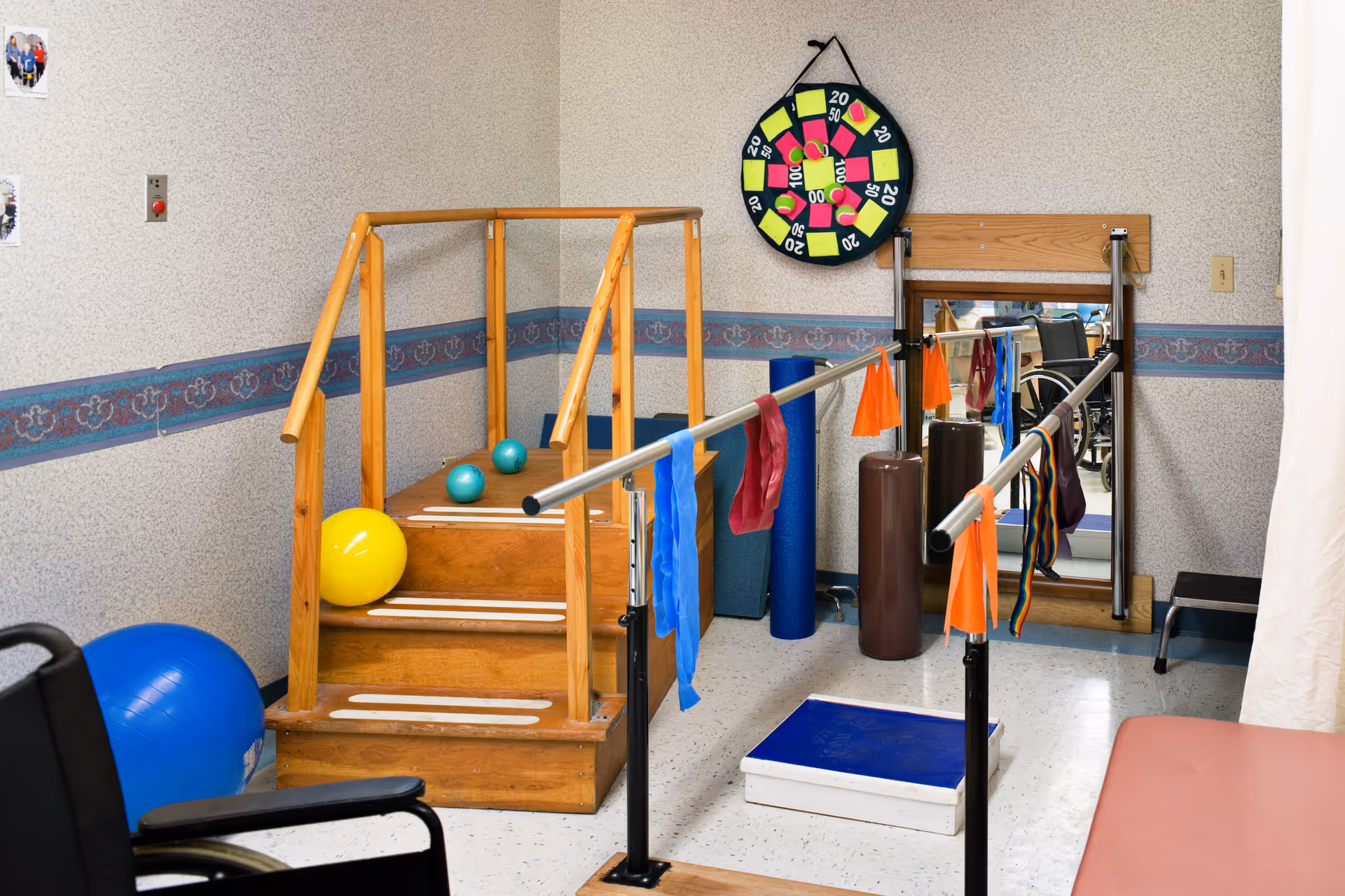 A physical therapy room with wooden stairs equipped with handrails, colorful exercise balls, resistance bands hanging on parallel bars, a mirror on the wall, a dartboard, and a wheelchair partially visible in the foreground.