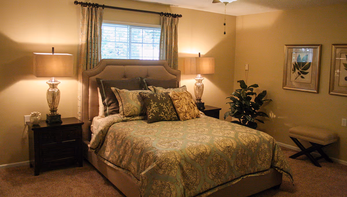 A cozy bedroom with a neatly made bed featuring patterned bedding and multiple decorative pillows. Two bedside tables each hold a lamp with a beige shade. A window with curtains is above the bed, and there are two framed botanical prints on the wall. A small bench and a potted plant are also visible in the room.