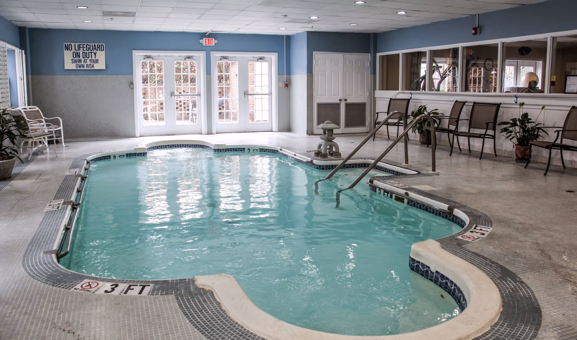 Indoor swimming pool with handrails, seating along the wall, and double exit doors at the far end.