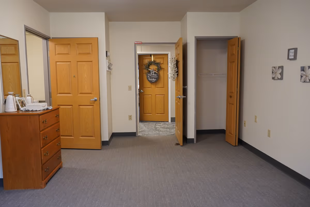 Interior view of a room with beige walls and gray carpet. There are three wooden doors, one leading to a hallway with a door decorated with a wreath and a sign that says 'Home Sweet Home.' Another door opens to a closet with a wire shelf. A wooden dresser with decorative items and a mirror is on the left side. The walls have a few small framed decorations.