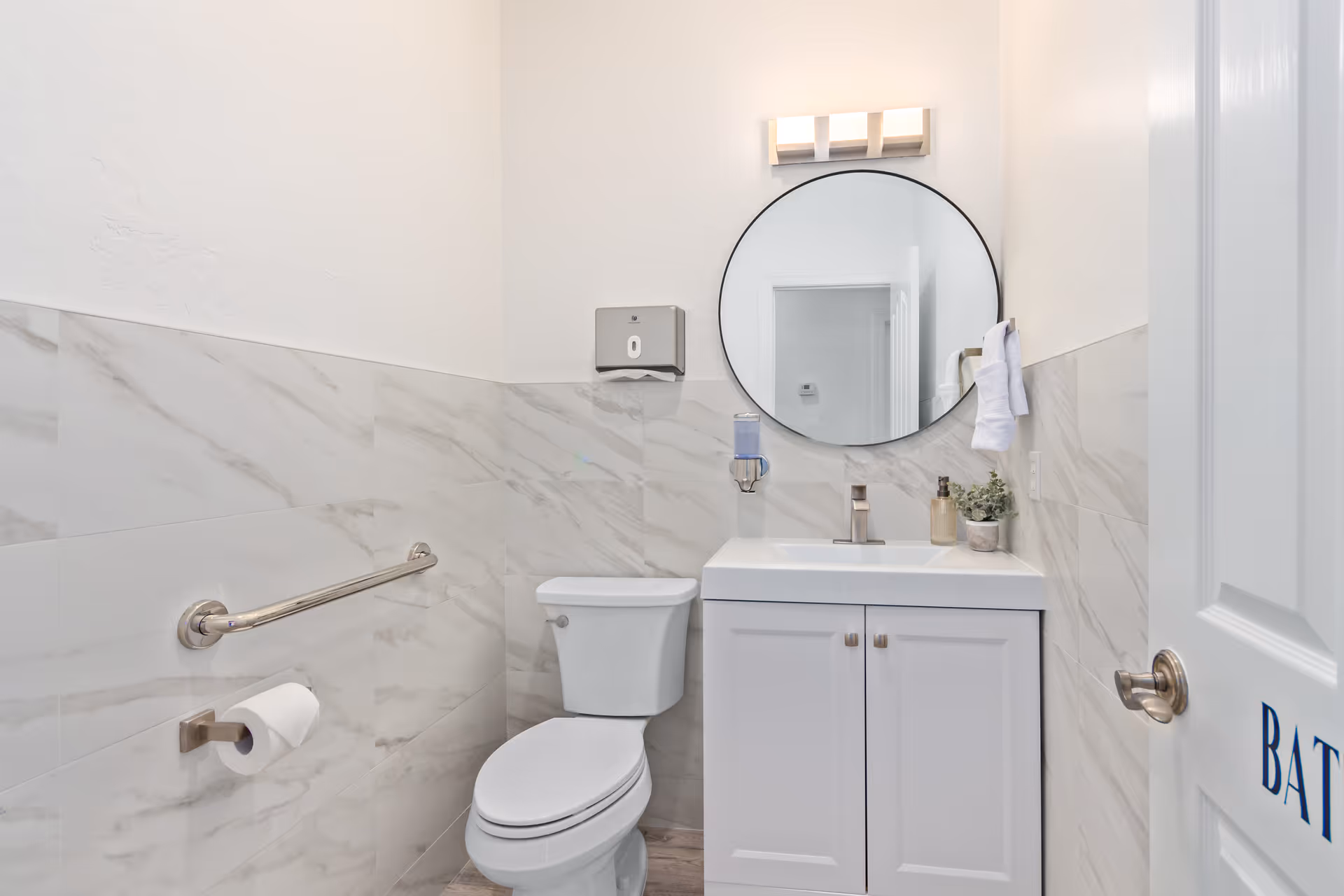 A clean and modern bathroom with white marble-patterned walls, a white toilet, a white vanity with a sink, a round mirror above the sink, a wall-mounted soap dispenser, a towel ring with a white towel, and a grab bar next to the toilet. The door to the bathroom is partially open.