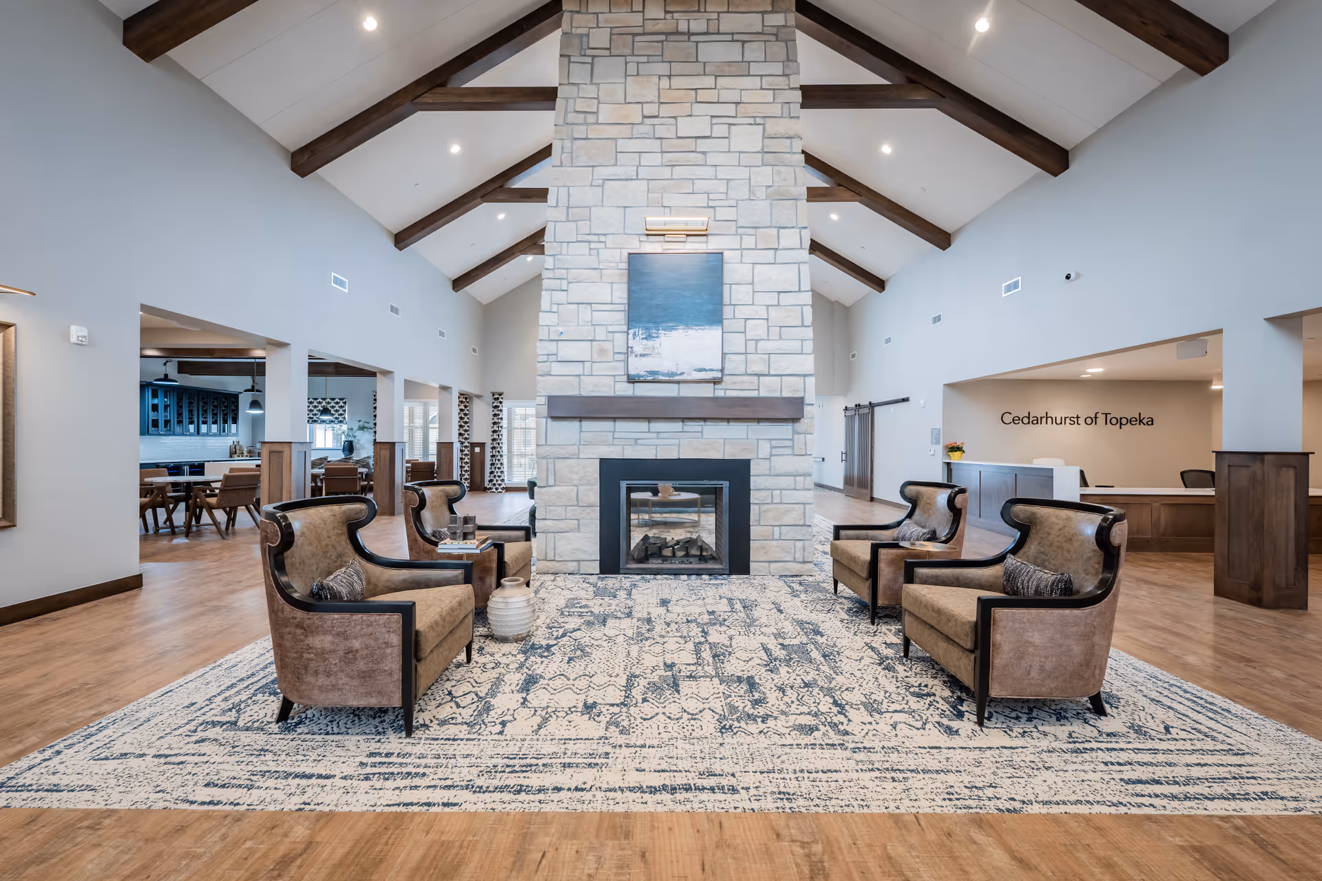 Spacious senior living facility common area with a high vaulted ceiling featuring exposed wooden beams, a large stone fireplace in the center, four upholstered armchairs arranged around the fireplace on a patterned area rug, and a reception desk with the sign 'Cedarhurst of Topeka' in the background.