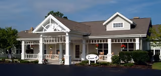 Exterior view of a single-story senior living community building with a covered entrance, white siding, and a gabled roof. There are hanging flower baskets and shrubs around the entrance, with a sign that reads 'The Wyngate'.