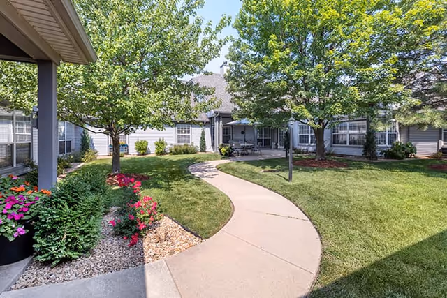 A landscaped outdoor courtyard area at Brookdale Tallgrass featuring a curved concrete walkway, green grass, trees, colorful flowers, and a covered seating area attached to a building with multiple windows.