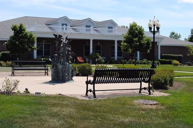 Outdoor courtyard area with black metal benches surrounding a decorative fountain sculpture. The background shows a single-story brick building with white trim, windows, and a porch with chairs. There are green lawns, bushes, and trees under a blue sky with some clouds.