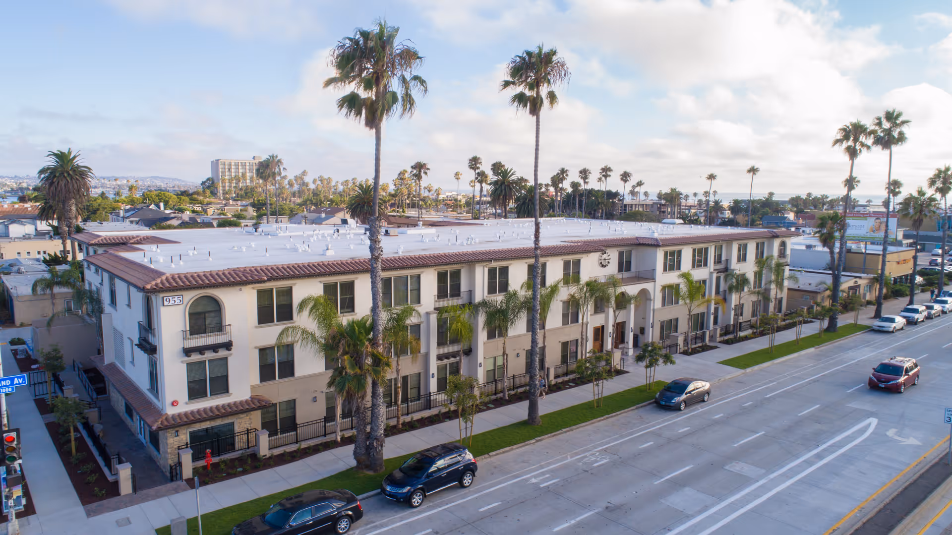 Front façade of a three-story Mediterranean-style senior living building with palm trees lining the sidewalk and cars on the street.