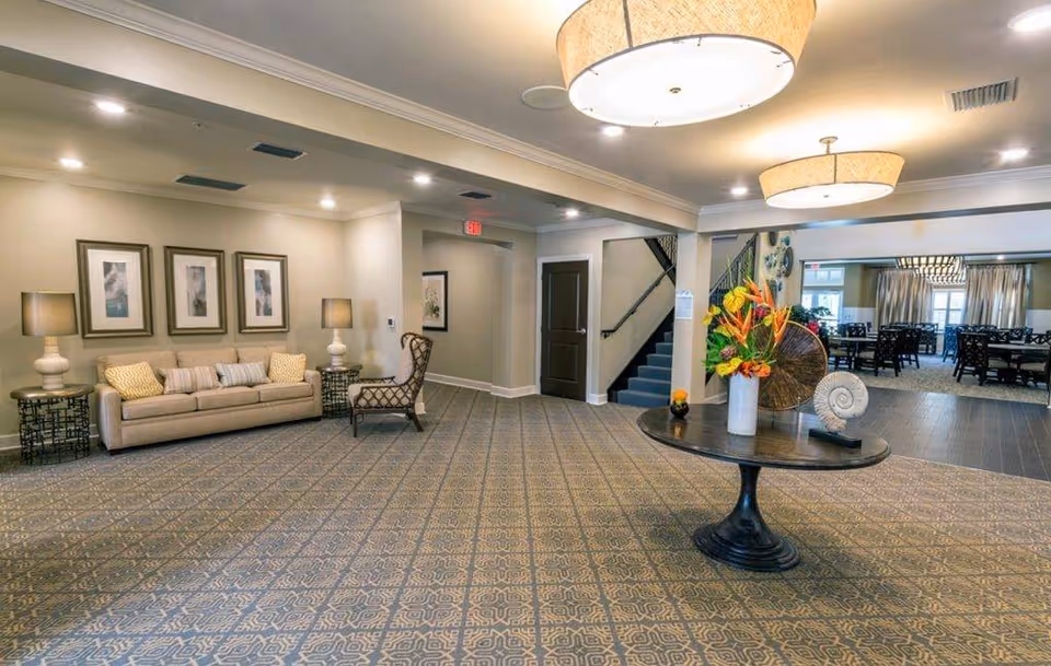 A spacious and well-lit interior common area with a patterned carpet, a round table with a floral arrangement and decorative items in the center, a beige sofa with cushions, two side tables with lamps, a single armchair, framed artwork on the wall, and a staircase leading to an upper floor. In the background, there is a dining area with multiple tables and chairs, large windows, and curtains.