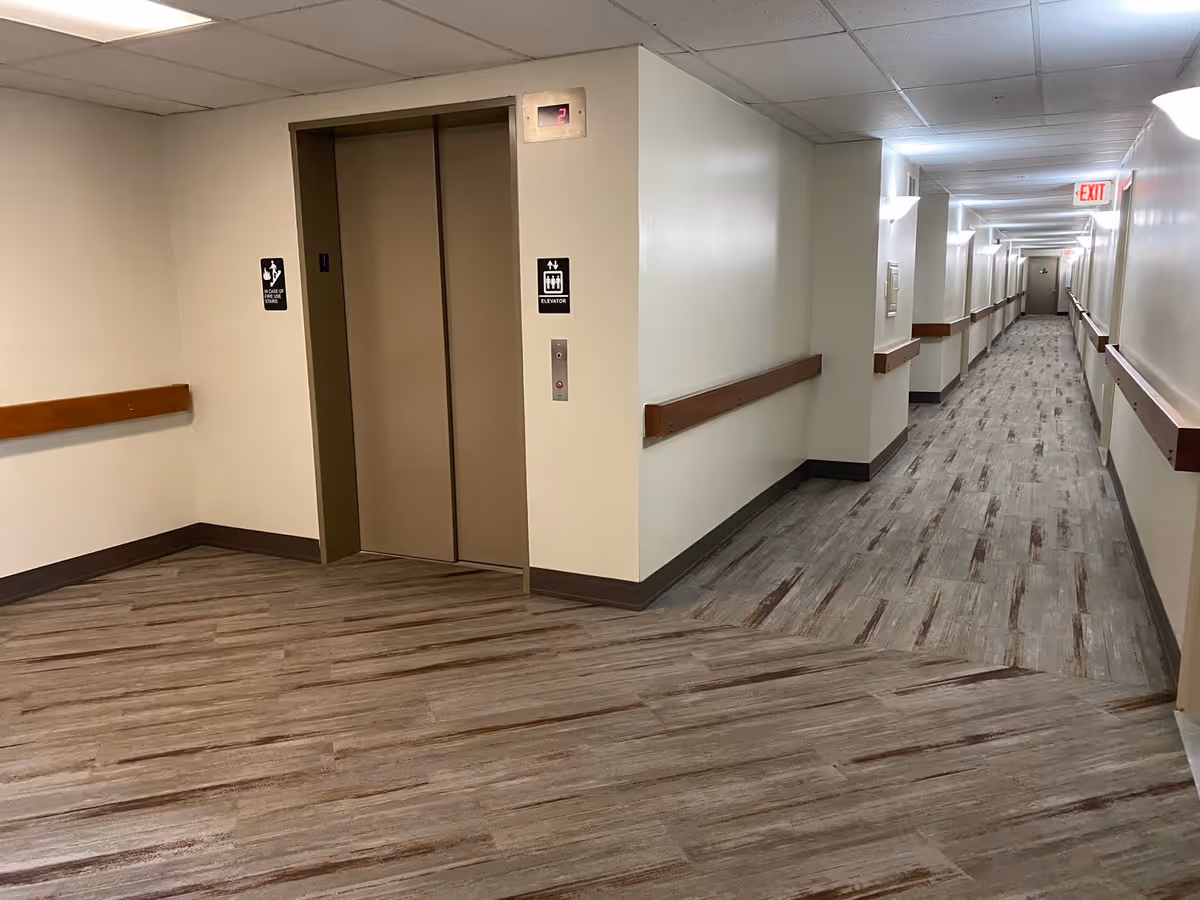 Interior hallway of Shelby Manor Senior Living featuring a closed elevator with beige doors on the left side and a long corridor extending to the right with handrails along the walls and an exit sign at the far end.