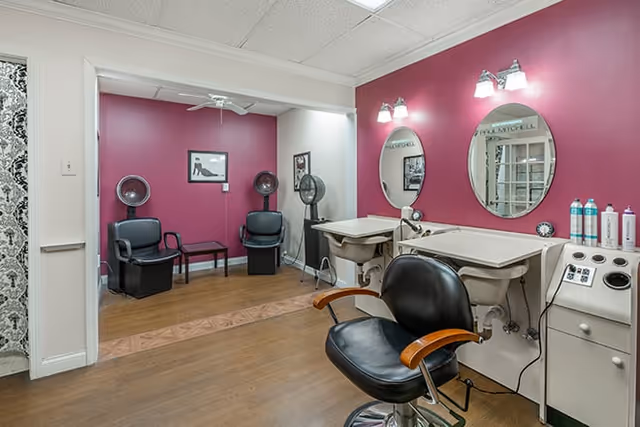 Interior view of a hair salon area in a senior living facility with two black salon chairs in front of sinks and mirrors on a magenta wall. In the background, there are two black chairs with hair dryers and a small table between them. The floor is wooden and the ceiling is white with recessed lighting.