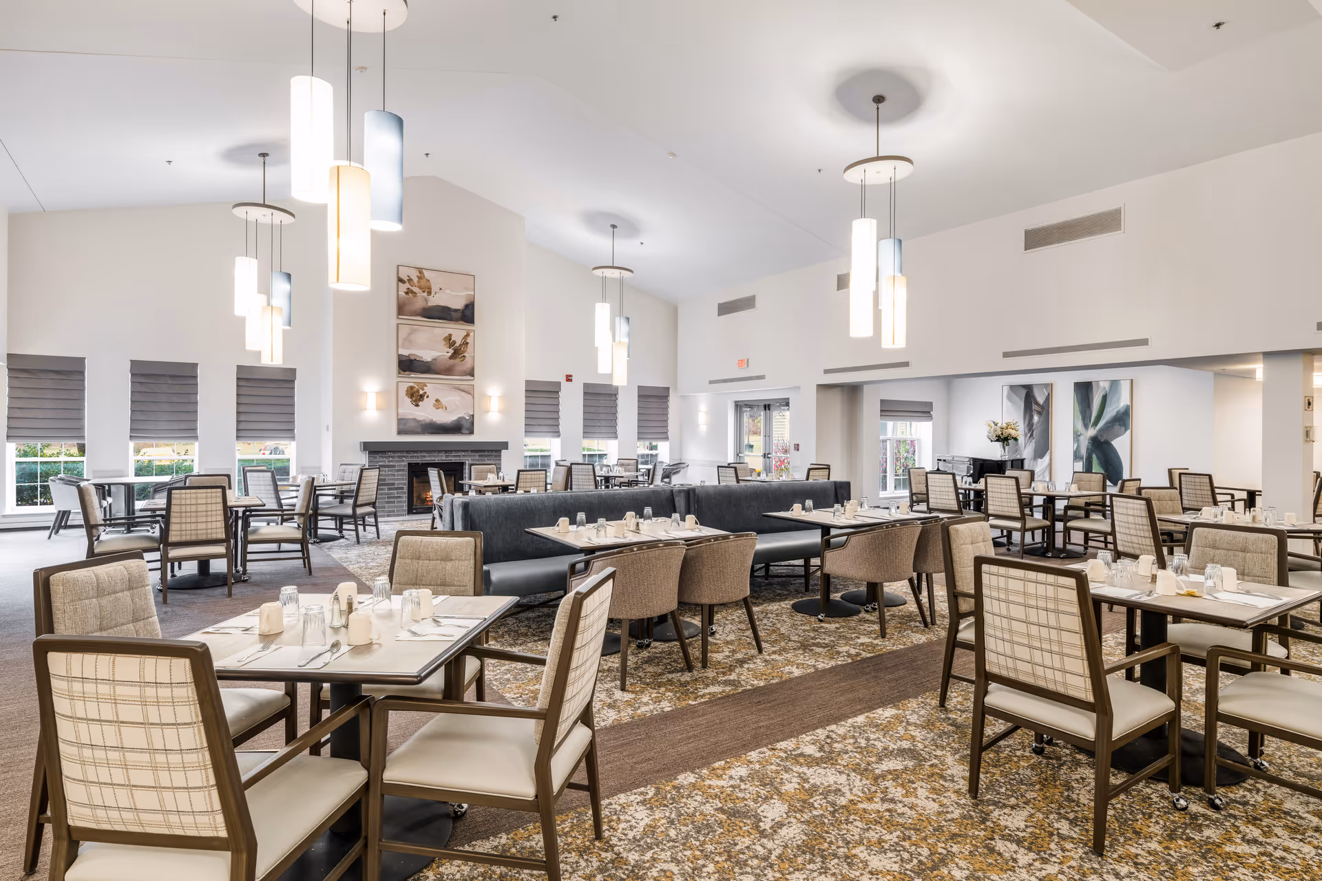 A spacious and well-lit dining room in a senior living community with multiple tables and chairs arranged neatly. The room features modern pendant lights hanging from a high ceiling, large windows with gray blinds, a fireplace with decorative artwork above it, and a patterned carpet floor. Tables are set with glasses, napkins, and utensils.
