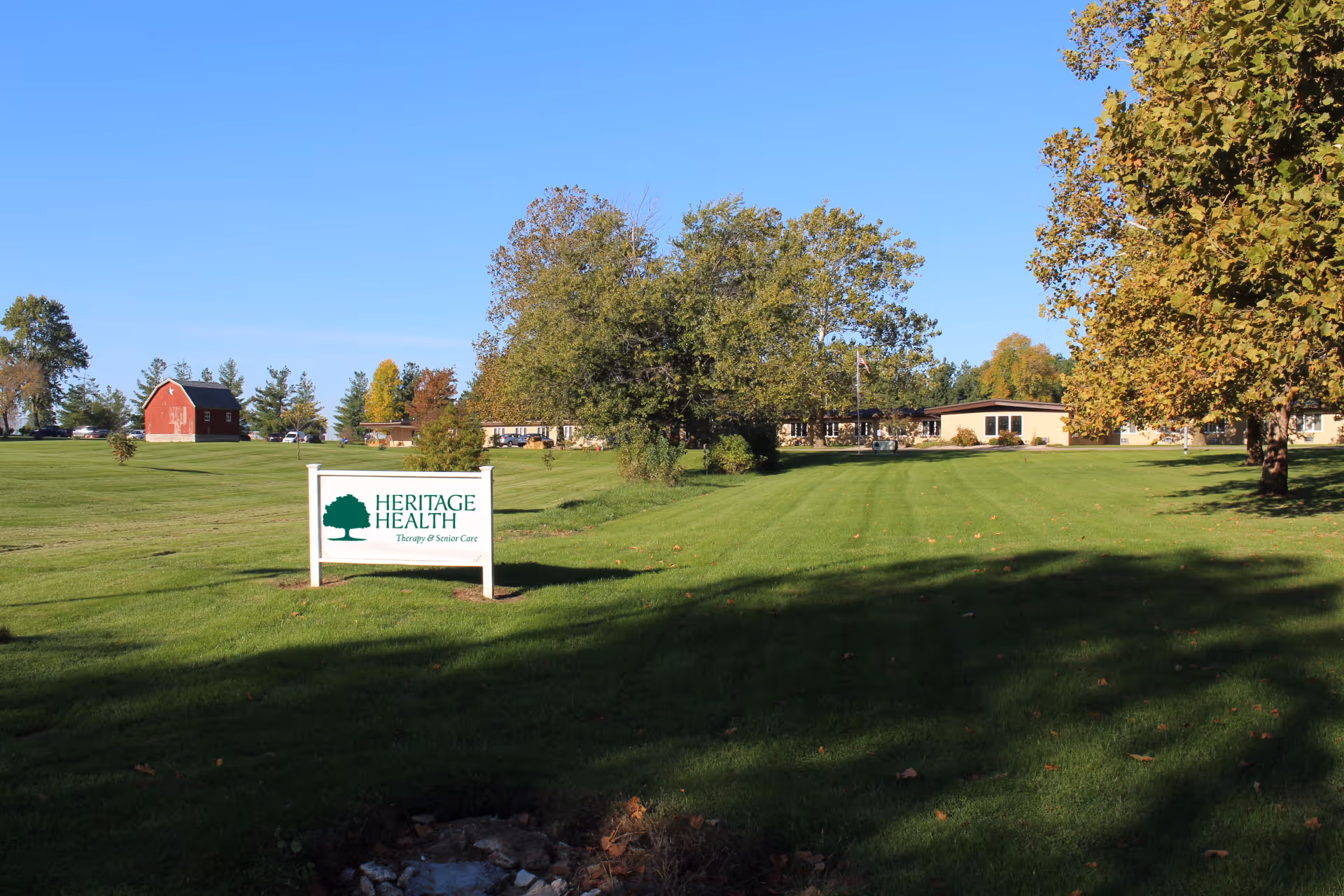 A large green lawn with a white sign that reads 'HERITAGE HEALTH Therapy & Senior Care' in front of a single-story building surrounded by trees under a clear blue sky.