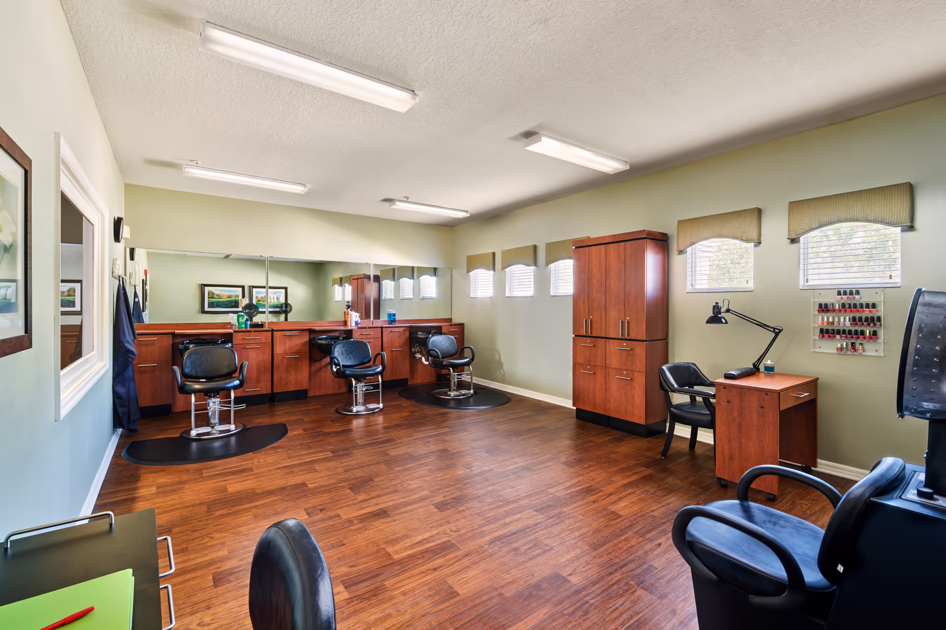 A spacious salon room with wooden flooring and light green walls. The room features three black salon chairs in front of a large mirror with wooden cabinetry underneath. There is a wooden cabinet and a small desk with a black chair and a lamp on the right side. Nail polish bottles are displayed on the wall near the desk. The room is well-lit with ceiling lights and natural light coming through small windows with beige valances.