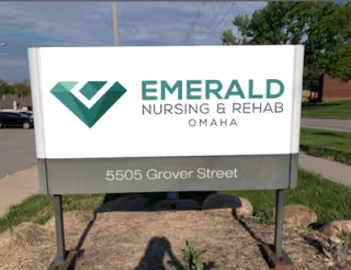 Outdoor sign for Emerald Nursing & Rehab Omaha with logo and the address "5505 Grover Street" on a grassy roadside.