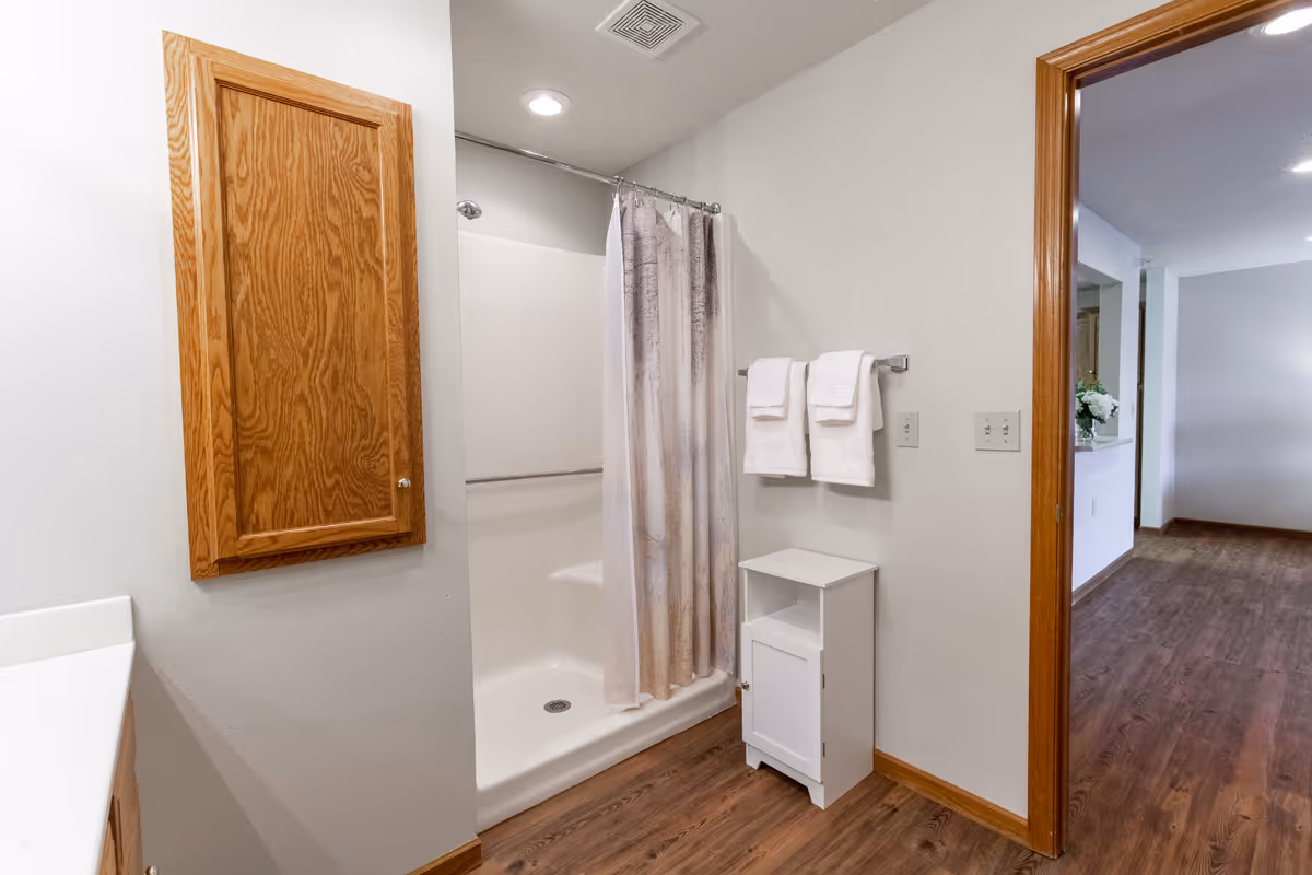 Bright bathroom featuring a shower with a curtain, a wooden medicine cabinet, a towel rack with white towels, and a small white storage cabinet opening to a wood-floored hallway.