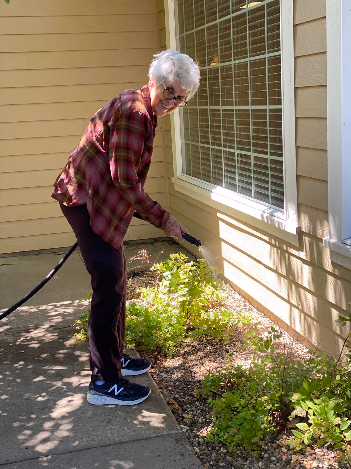 An elderly woman with short white hair and glasses is watering plants in a garden bed next to a beige house with a large window. She is wearing a red plaid shirt, dark pants, and black New Balance sneakers, standing on a concrete pathway.