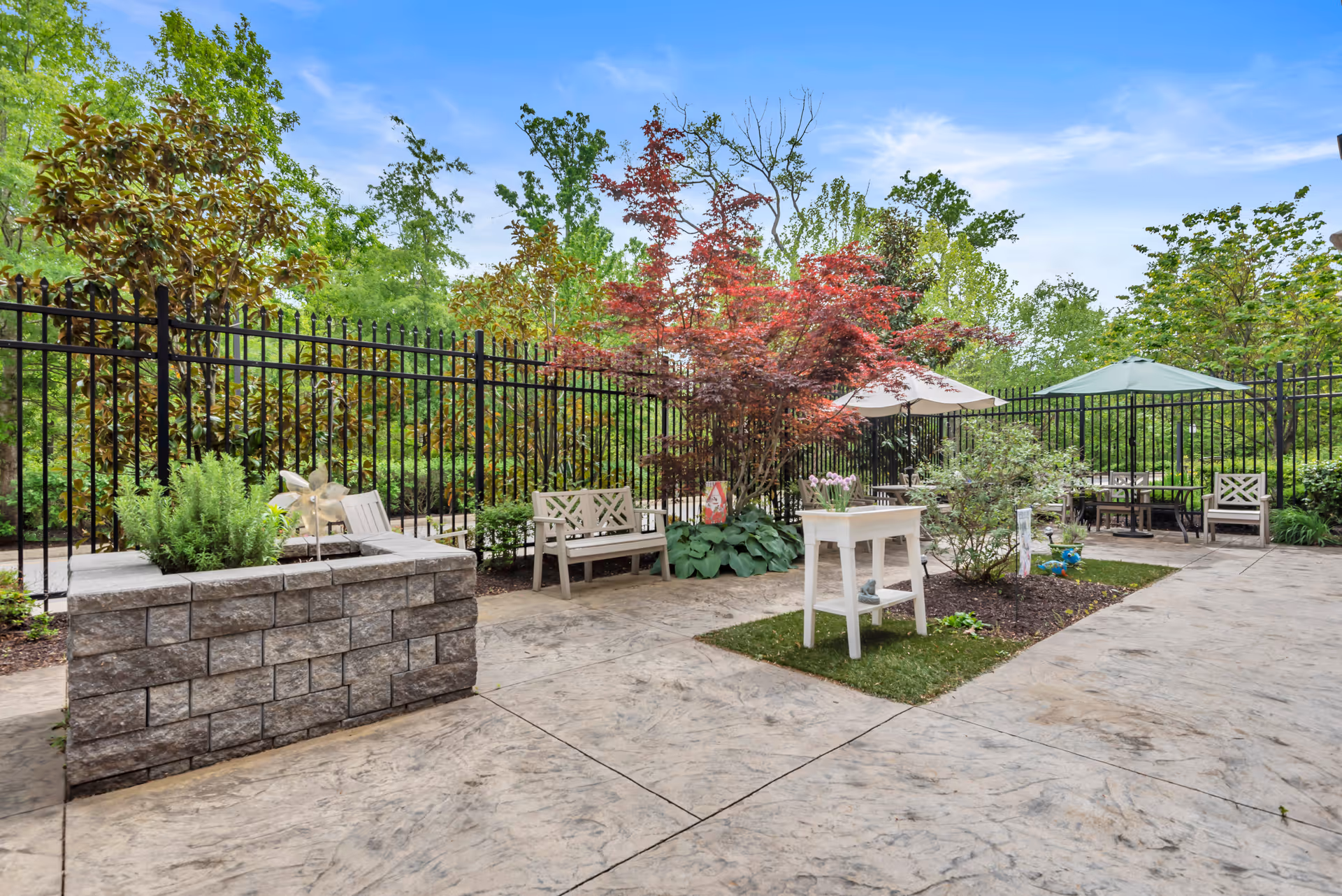 Enclosed outdoor patio with benches, umbrellas, planters, and ornamental trees against a black metal fence.