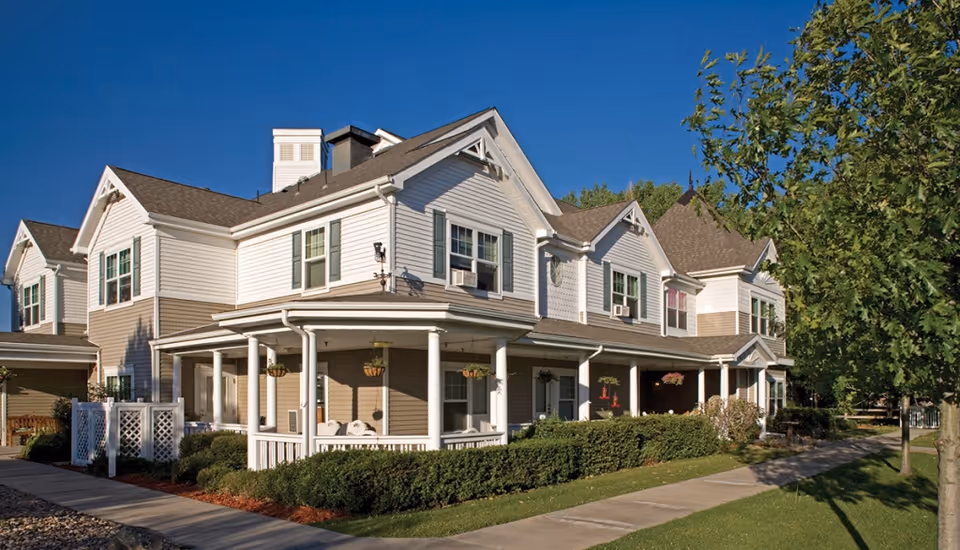 Exterior view of a two-story assisted living facility building with white and beige siding, multiple windows, a covered porch with hanging flower baskets, surrounded by green shrubs and trees under a clear blue sky.