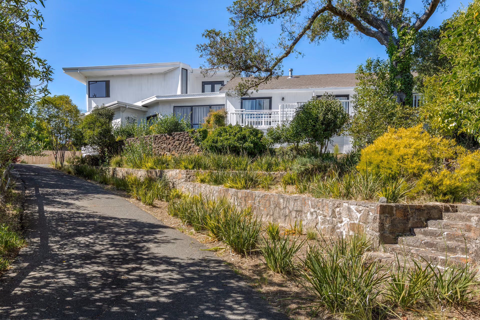 A white multi-level building with large windows and a balcony, surrounded by lush greenery and trees under a clear blue sky. A paved driveway leads up to the building, with stone retaining walls and steps on the right side.