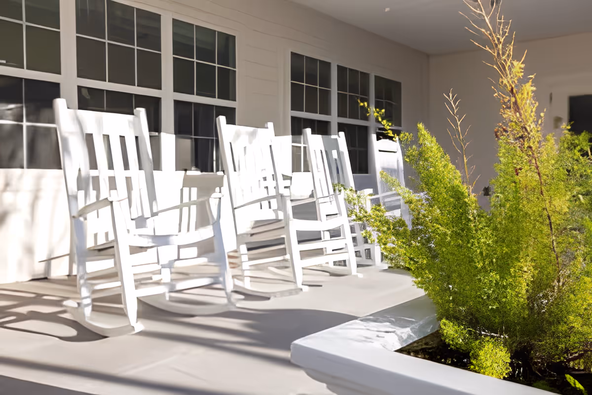 A sunny porch with several white wooden rocking chairs in front of large windows and green shrubs.
