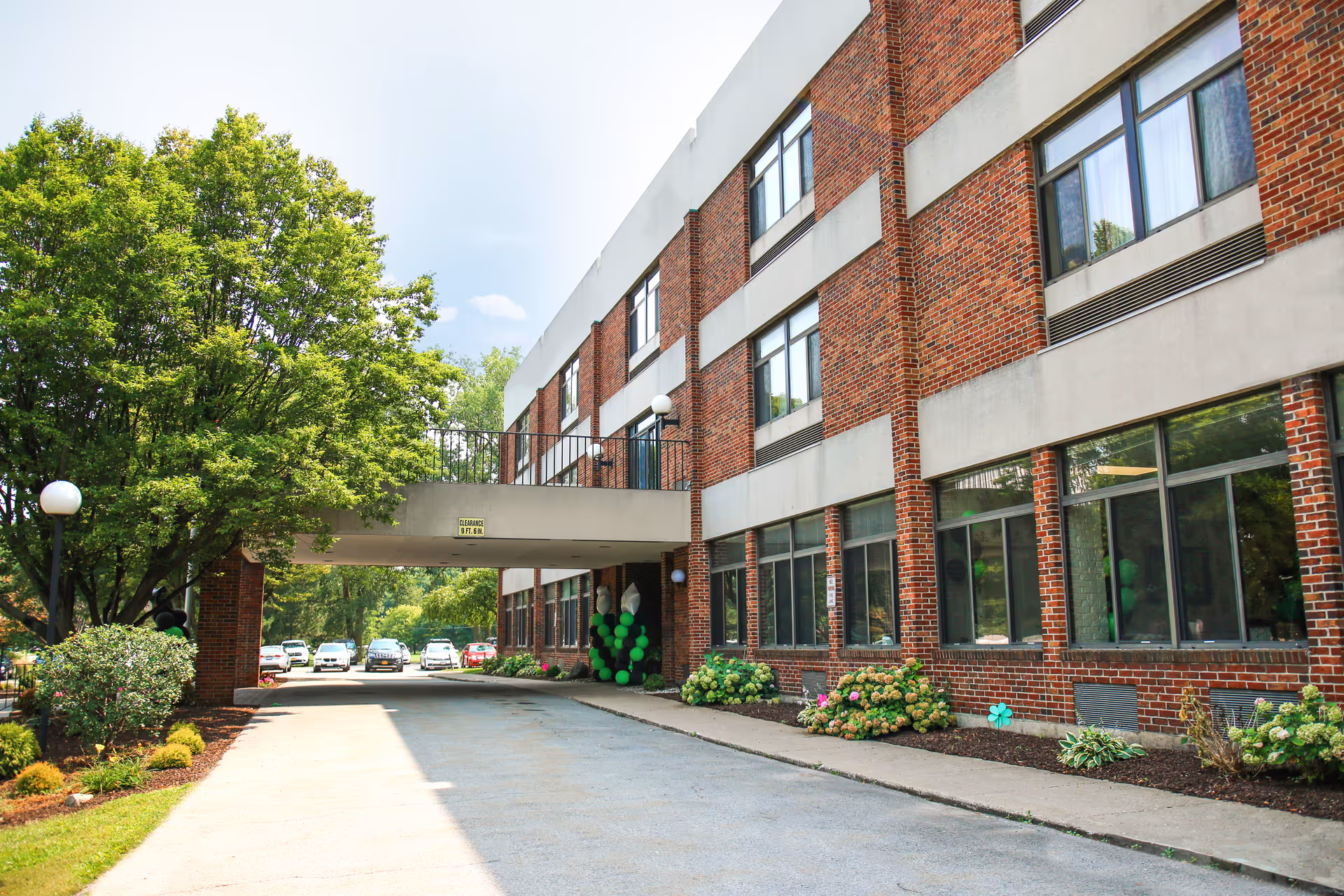 Exterior view of a three-story brick building with large windows, a covered driveway, and green trees and shrubs surrounding the area. Several cars are parked in the background under a partly cloudy sky.
