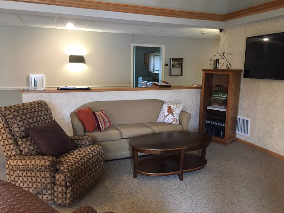 A cozy living room area with a beige sofa adorned with three decorative pillows, a patterned armchair with a brown cushion, and a wooden oval coffee table. There is a wooden shelving unit with decorative items and a flat-screen TV mounted on the wall. The room has beige walls with a wooden trim and carpeted floor. A doorway in the background leads to another room with kitchen cabinets visible.