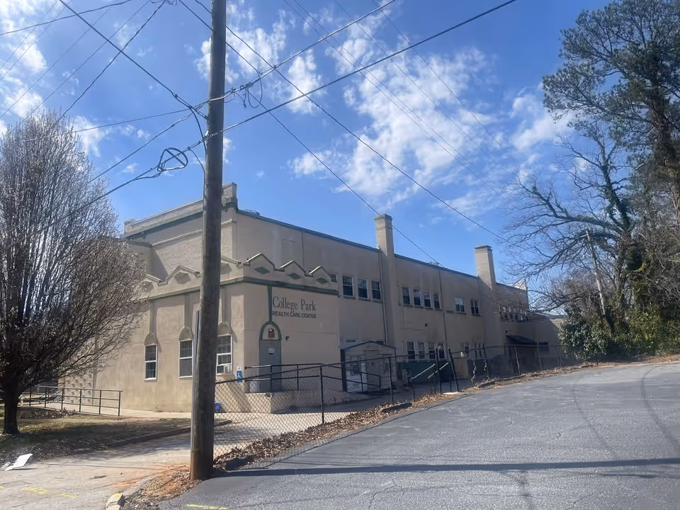 Two-story beige health care building with a fenced entrance, utility pole, and trees under a blue sky.