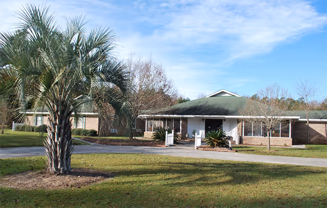 Single-story assisted living facility building with a green roof, surrounded by grass and trees, including a large palm tree in the foreground under a clear blue sky.