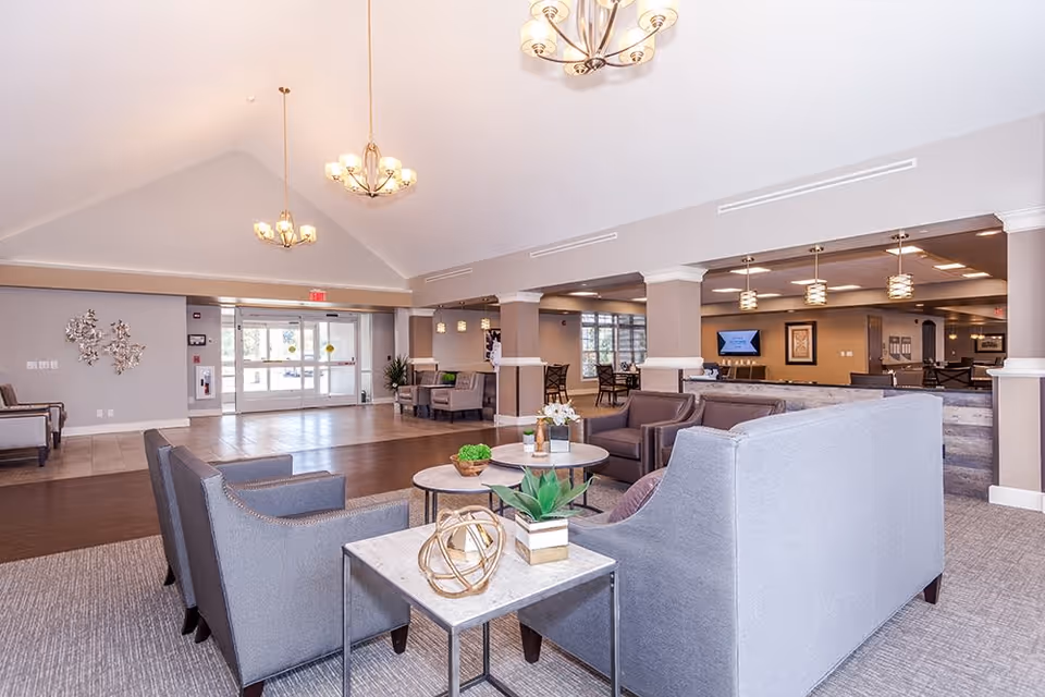 A spacious and well-lit assisted living facility common area with modern gray sofas and armchairs arranged around circular coffee tables. The room features high vaulted ceilings with elegant chandeliers, a mix of carpet and tile flooring, and large glass entrance doors in the background. The area includes decorative plants and wall art, with additional seating and a television visible further inside the space.