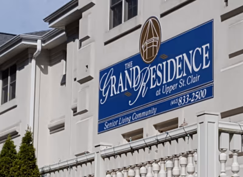 Exterior view of a senior living community building with a large blue sign that reads 'The Grand Residence at Upper St. Clair Senior Living Community' along with a phone number.