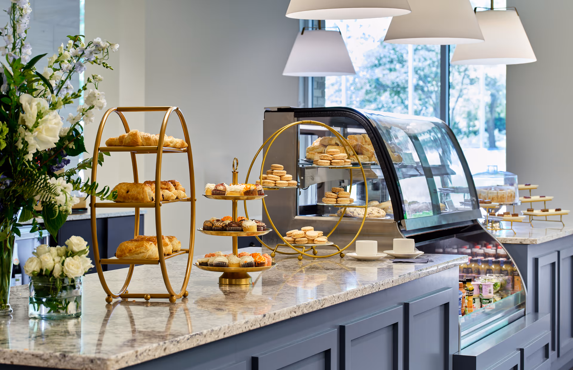 A countertop display in a dining area featuring an assortment of pastries and desserts on tiered golden stands, with a glass refrigerated case containing more baked goods and beverages. There are also white cups and saucers on the counter, and a vase with white and green flowers to the left. Large windows in the background let in natural light.