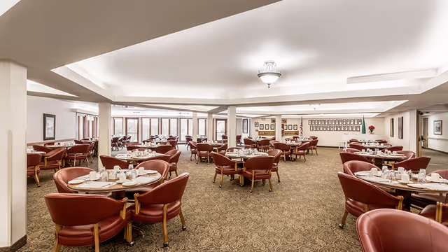 Spacious dining room with multiple round tables set with white tableware and surrounded by red cushioned chairs. Large windows along one side let in natural light, and framed pictures decorate the walls.