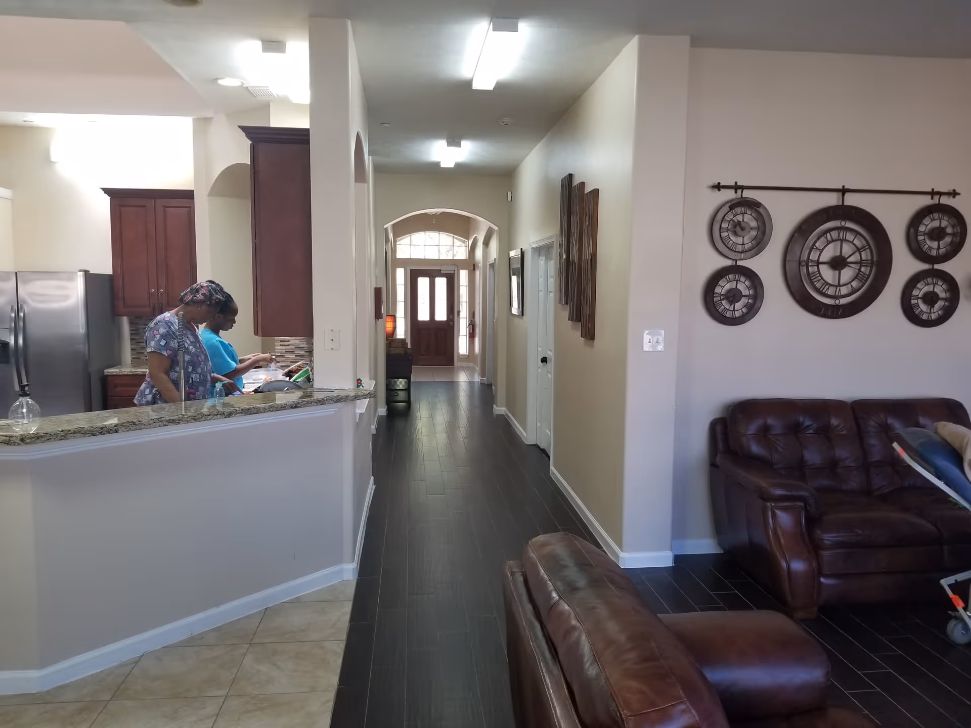 Interior view of a senior living facility showing a kitchen area on the left with two staff members working, a hallway leading to a wooden front door with glass panels, and a living room area on the right with brown leather sofas and a decorative wall clock with multiple circular faces.