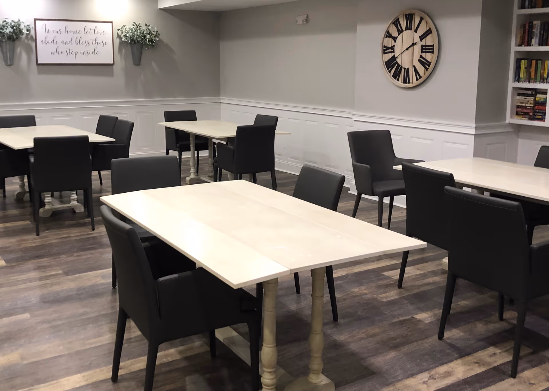 A dining room with multiple white tables and black chairs arranged neatly on a wooden floor. The walls are light gray with white wainscoting, decorated with a large round clock, two small wall plants, a framed sign with cursive writing, and a bookshelf filled with books.