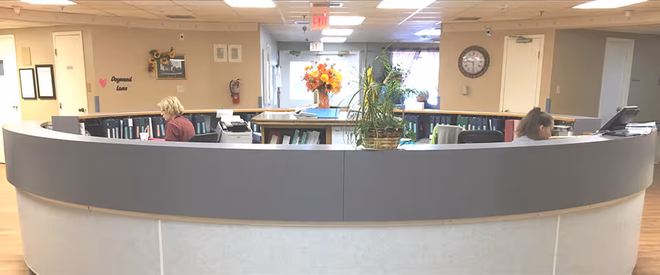 Reception area with a curved desk and two staff members working behind it. The desk has office supplies, binders, and a potted plant with orange flowers. The background shows a hallway with a clock on the wall and some framed pictures.