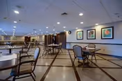 Spacious communal dining room with round tables and chairs, tiled floor, drop ceiling lights, and framed artwork on the walls.