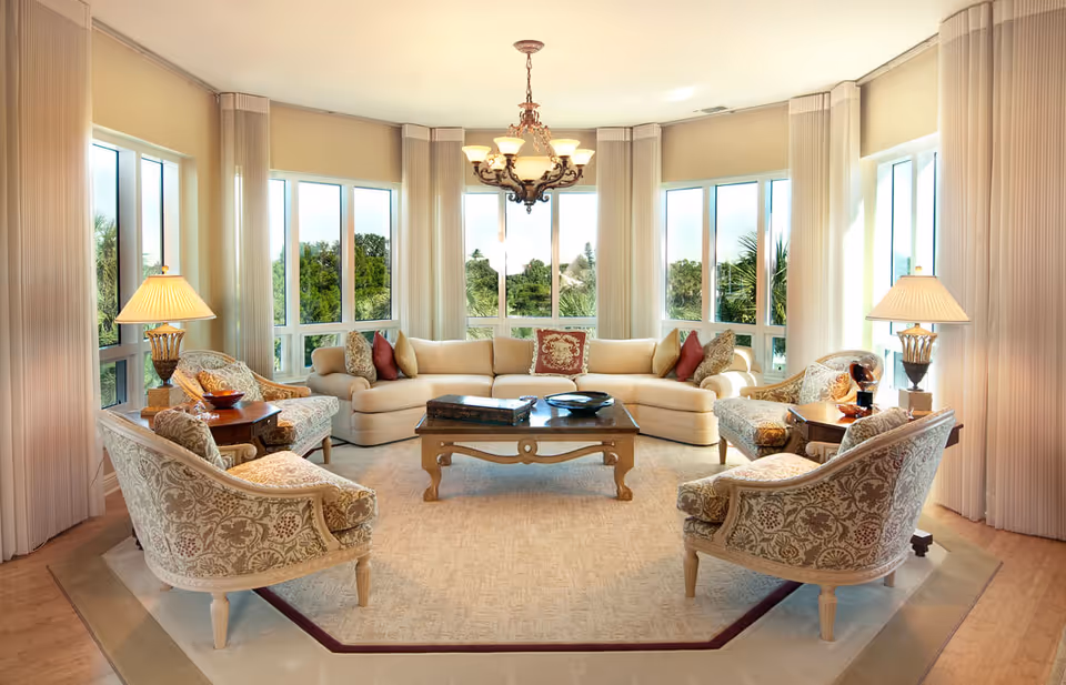 A bright and elegant living room with large windows offering a view of greenery outside. The room features a beige sectional sofa adorned with decorative pillows, four patterned armchairs arranged around a wooden coffee table, two side tables with lamps, and a chandelier hanging from the ceiling.