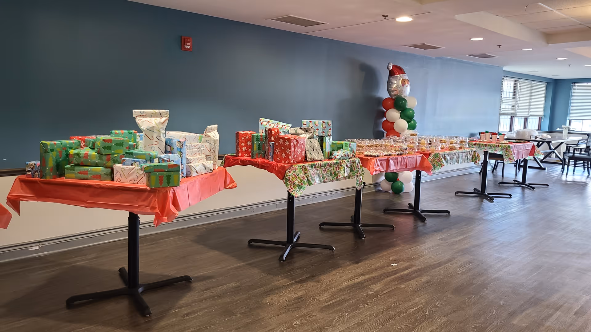 A long row of tables covered with festive holiday tablecloths displaying wrapped Christmas presents and trays of baked goods in a spacious room with blue walls and large windows. A balloon decoration shaped like a snowman wearing a Santa hat is visible near the wall.