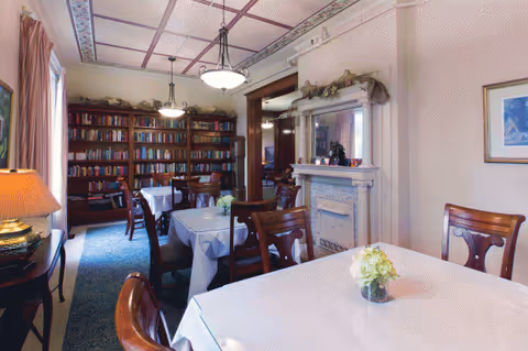 A dining room with white-clothed tables, flowers, chairs, a fireplace, and a bookshelf.
