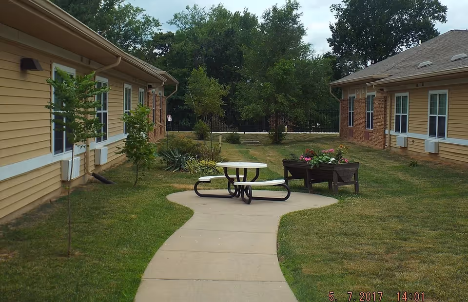 Outdoor courtyard area between two single-story buildings with beige siding and brick accents. There is a curved concrete walkway leading to a round picnic table with attached benches and a wooden planter box with flowers. Trees and shrubs are planted along the buildings, and a black metal fence is visible in the background with dense green trees beyond.