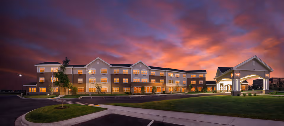 Exterior view of Primrose Retirement Community of Pleasant Prairie at dusk, showing a large three-story building with many lit windows, a covered entrance, well-maintained lawn, and a dramatic colorful sky with clouds.