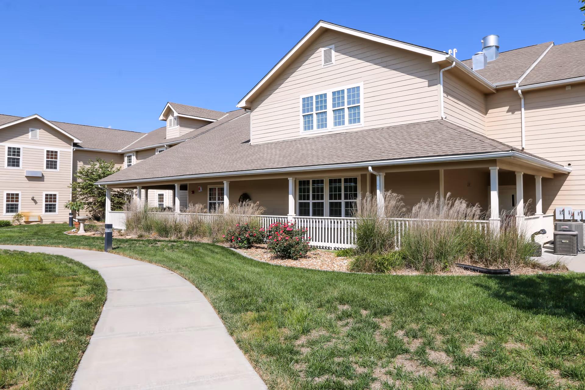 Exterior view of a beige two-story senior living facility building with a covered porch, white railing, and landscaped garden with grass, bushes, and flowers. A curved concrete walkway leads to the entrance under a clear blue sky.
