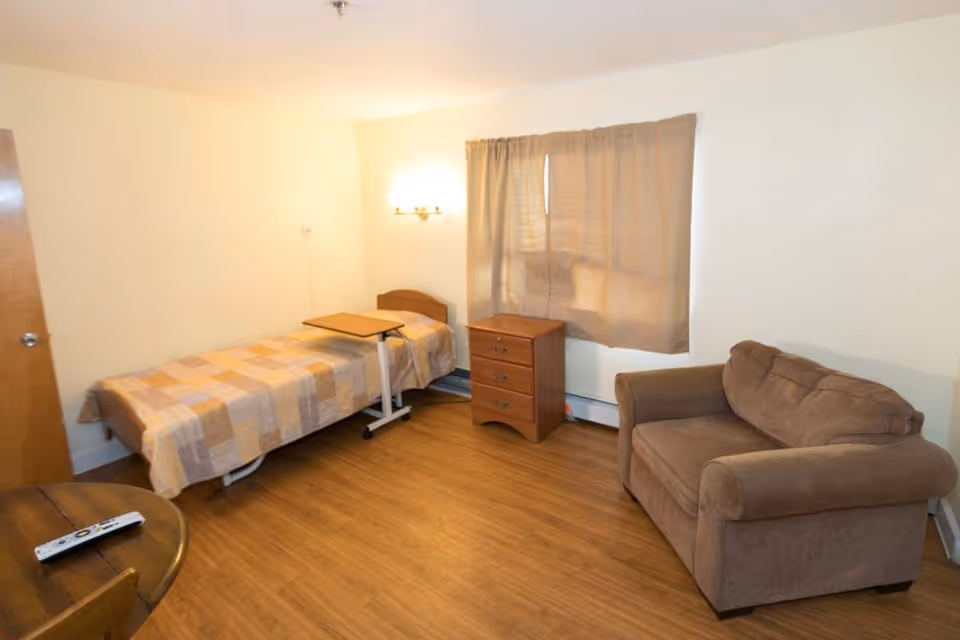A small room with a single bed covered with a checkered beige and light brown blanket, a wooden bedside table with three drawers, a brown upholstered armchair, a round wooden table with a TV remote on it, and a window with beige curtains. The floor is wooden and the walls are painted light yellow.