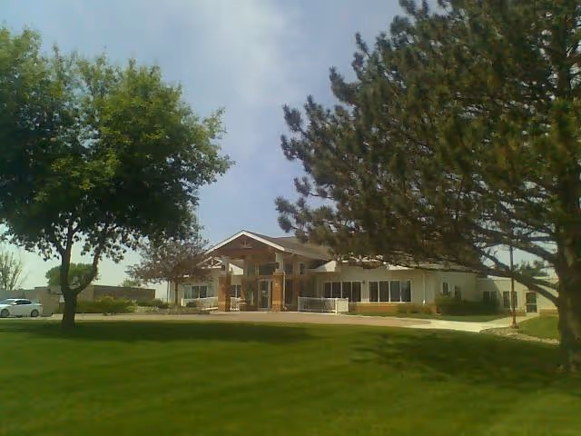 Exterior view of a single-story building with a peaked roof entrance, surrounded by green grass and trees under a partly cloudy sky.