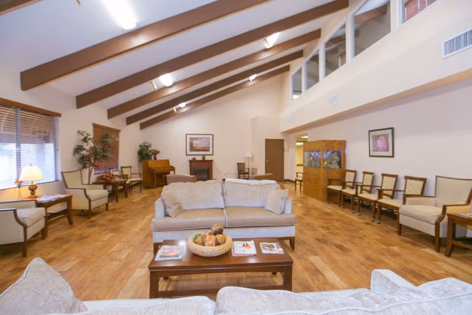 A spacious living room area in Sherwood Village Assisted Living & Memory Care featuring beige sofas with cushions, a wooden coffee table with magazines and a decorative bowl, multiple chairs lined up against the walls, a piano in the corner, framed artwork on the walls, large windows with blinds, wooden ceiling beams, and warm lighting.