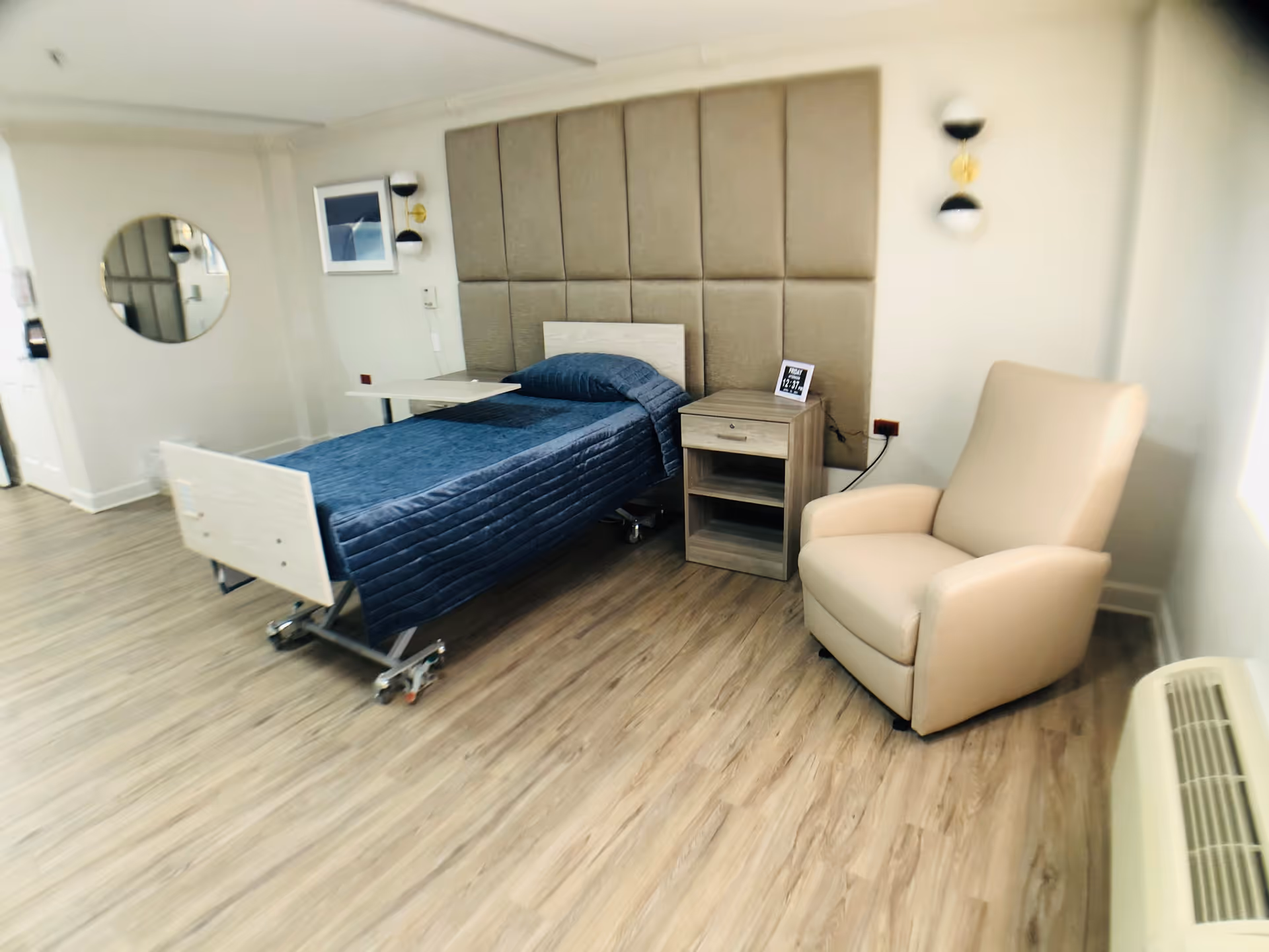 A senior living bedroom with an adjustable single bed with blue bedding, a padded headboard, a nightstand, and a beige recliner.
